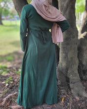 Load image into Gallery viewer, Woman wearing a green maxi dress tied at the waist and tan hijab standing next to a tree in a park.