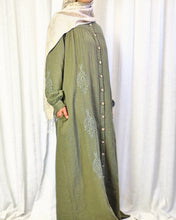 Load image into Gallery viewer, Side view of woman wearing a long khaki dress with buttons and embroidery against a white curtain background