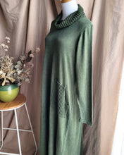 Load image into Gallery viewer, A khaki green knit cowl neck dress with a ribbed pocket detail, displayed on a mannequin torso against a beige curtain backdrop with a small wooden stool and dried flowers on the left.