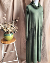 Load image into Gallery viewer, A khaki green knit maxi dress with a ribbed cowl neck and two large ribbed patch pockets, displayed on a mannequin next to a small wooden stool with a green ceramic vase of dried flowers against a beige fabric backdrop.
