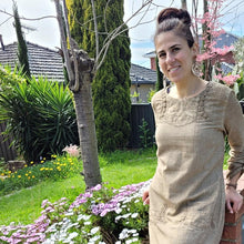 Load image into Gallery viewer, Sandy-tunic-dress-with-tab-button-long-sleeves-jewel-neckline-applique-detail-on-front-yoke-and-pockets-and-string-tie-waist-in-sand-next-to-brick-wall-close-up