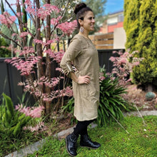 Load image into Gallery viewer, Sandy-tunic-dress-with-tab-button-long-sleeves-jewel-neckline-applique-detail-on-front-yoke-and-pockets-and-string-tie-waist-in-sand-next-to-pink-tree-side-view