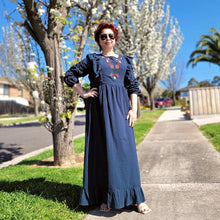 Load image into Gallery viewer, ginger-long-sleeve-embroidered-maxi-dress-with-ruffled-trim-over-shoulders-cinched-waistline-with-sash-tie-and-ruffled-hem-in-teal-next-to-trees-in-bloom