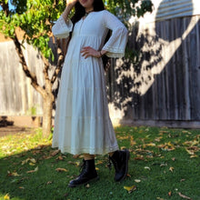 Load image into Gallery viewer, Sharon_tiered_midi_dress_in_cream_with_Vneckline_fitted_buttoned_bust_tiered_skirt_with_lace_trim_at_hem_and_bell_sleeves_with_lace_trim_with_boots_front_of_garden_tree