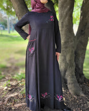 Load image into Gallery viewer, Woman in a long black dress with floral embroidery and trim on front pocket and neckline standing next to a tree in a park.
