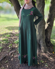 Load image into Gallery viewer, Woman in a green dress with floral embroidery detail and hijab standing under a tree in a park.
