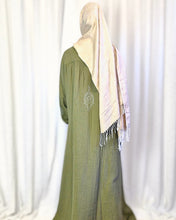 Load image into Gallery viewer, Back view of woman wearing a long khaki abaya/dress with a beige headscarf showing embroidery detail on the back against a white curtain background
