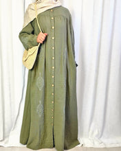 Load image into Gallery viewer, Woman wearing a long abaya dress with buttons and embroidery against a white curtain background
