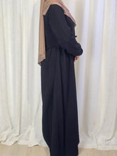 Load image into Gallery viewer, Side view of woman wearing a long black dress with a beige hijab against a white curtain background

