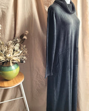 Load image into Gallery viewer, A charcoal gray knit cowl-neck maxi dress with long sleeves and large ribbed patch pockets, displayed on a mannequin against a beige fabric backdrop next to a small wooden stool with a green vase of dried flowers.
