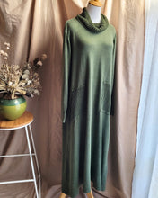 Load image into Gallery viewer, A khaki green knit cowl-neck dress displayed on a mannequin torso, featuring ribbed knit details on the pockets and cowl neck. The dress is styled against a backdrop of beige and white curtains, with a small wooden stool and a green ceramic pot with dried flowers on the left.
