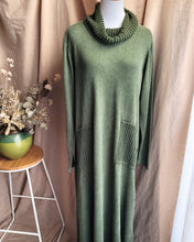 Load image into Gallery viewer, An khaki green knit maxi dress with a ribbed cowl neck and two large ribbed patch pockets, displayed on a white mannequin torso against a beige curtain backdrop with a small wooden stool and dried flowers on the left.
