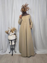 Load image into Gallery viewer, Woman wearing beige modest cotton muslin maxi dress with pintuck detail on bodice and sleeves, long sleeve flowing silhouette, standing against white curtain and a large ornamental vase with dried flowers next to her.
