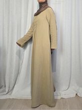 Load image into Gallery viewer, Side view of woman wearing beige modest cotton muslin maxi dress with flowing silhouette and long sleeves, standing against a white curtain.
