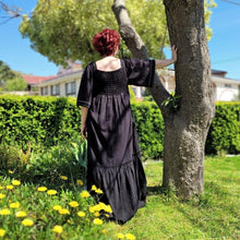 Load image into Gallery viewer, manolya-shirred-maxi-dress-with-bell-sleeves-square-neckline-empire-waist-skirt-with-ruffled-hem-in-black-outdoor-back-view-image-next-to-tree-trunk
