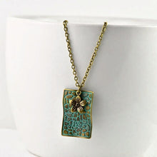 Load image into Gallery viewer, vintage-style-bronze-rectangle-pendant-necklace-with-verdigris-patina-finish-and-small-flower-charm-hanging-from-while-bowl
