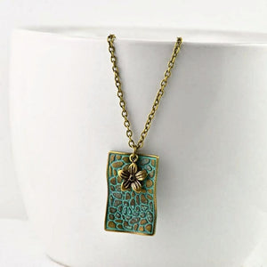 vintage-style-bronze-rectangle-pendant-necklace-with-verdigris-patina-finish-and-small-flower-charm-hanging-from-while-bowl